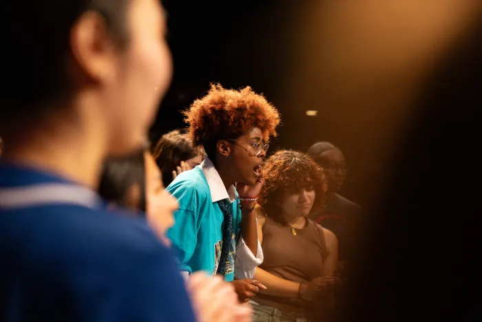 Students signing a cappella, one student particularly in focus, backlit by stage lights