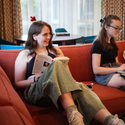 A student sitting on a sofa reading a book and laughing with someone off-screen.